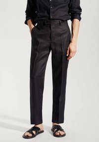 Man wearing black tailored pants, black button-up shirt with sleeves rolled, and black open-toe sandals, standing with hands in pockets.