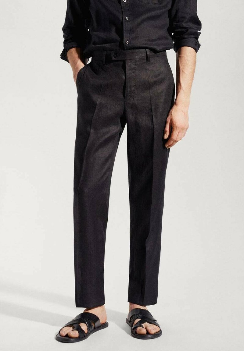 Man wearing black tailored pants, black button-up shirt with sleeves rolled, and black open-toe sandals, standing with hands in pockets.