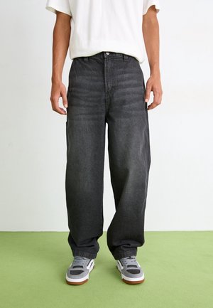 Personne portant un jean noir ample, un t-shirt blanc et des baskets grises, debout sur un sol vert contre un mur blanc uni.