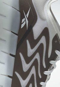 Baskets de sport marron et blanches avec une tige en maille texturée, un motif en zigzag et une semelle épaisse et segmentée, arborant des lacets blancs et un logo.