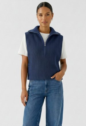 Frau trägt eine marineblaue, gerippte, ärmellose Weste mit Reißverschluss über einem weißen T-Shirt und blauen Jeans, mit einer Hand in der Tasche, stehend vor einem einfarbigen Hintergrund.