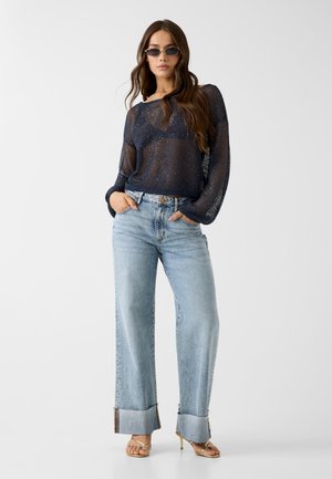 Vrouw draagt een zwart doorschijnend gebreid topje, lichtblauwe wijde jeans met omslag, gouden sandaaltjes met bandjes en donkere rechthoekige zonnebril, staat met handen in de zakken.