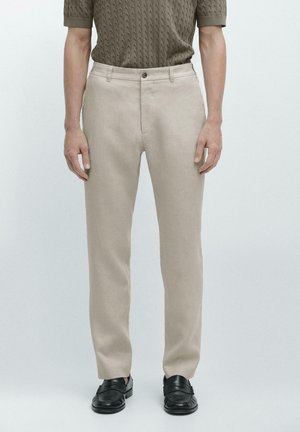 Uomo che indossa pantaloni beige a gamba dritta, mocassini in pelle neri e un maglione a maniche corte di colore verde oliva con trama, in piedi su sfondo bianco.