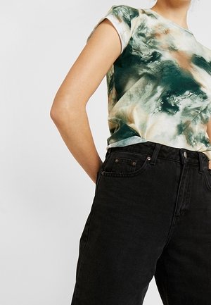 Person iført en grøn og beige tie-dye t-shirt stoppet ned i højtaljede sorte jeans med hænderne bag ryggen mod en ensfarvet baggrund.