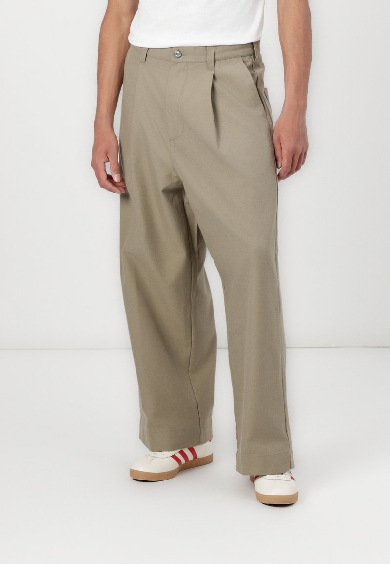 Homme portant un pantalon large beige, des baskets blanches avec des bandes rouges, et un t-shirt blanc, debout sur un sol blanc devant un fond uni.