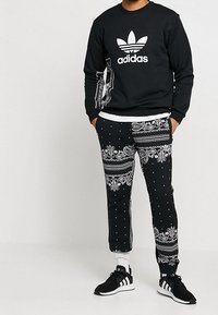 Černá mikina s bílým logem Adidas, v kombinaci s černými kalhotami s paisley a puntíkovanými vzory, doplněná černými sportovními botami.