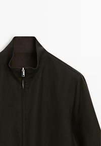 Veste noire avec un col montant et une fermeture éclair argentée partiellement fermée, présentée sur un fond blanc uni.