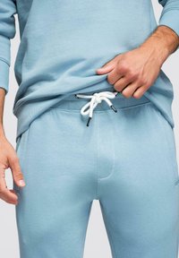 Homme portant un pantalon de jogging et un sweat-shirt bleu clair, soulevant le sweat-shirt pour montrer un cordon blanc noué en un nœud à la taille.