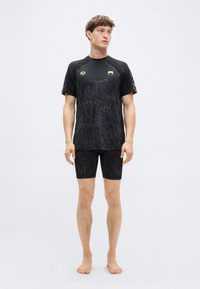 Μαύρο αθλητικό σετ με κοντομάνικο πουκάμισο με υφασμάτινη σχεδίαση και χρυσές λεπτομέρειες, συνδυασμένο με εφαρμοστά σορτς στο ίδιο χρωματικό σχήμα.