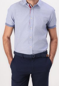 Camisa de manga corta azul claro con cuello abotonado, con un ribete en contraste de color rojo y azul marino. Combinada con pantalones azul marino y un cinturón trenzado.