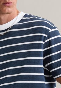 T-shirt rayé bleu marine avec des rayures blanches horizontales, texture côtelée et un col rond blanc. Présente un collier en chaîne argenté.