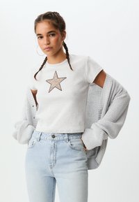 Svetlo siva majica s kratkimi rokavi z rahlo strukturo in grafiko zvezde. Ujemajoče se z visokim pasom svetlo modre kavbojke in udobnim svetlo sivim kardiganom.