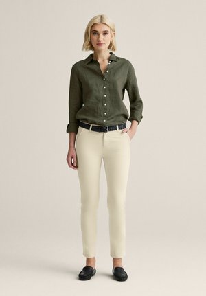 Nainen, jolla on lyhyet vaaleat hiukset ja päällään oliivinvihreä, napitettava paita, beiget housut, musta punottu vyö ja mustat slip-on kengät.