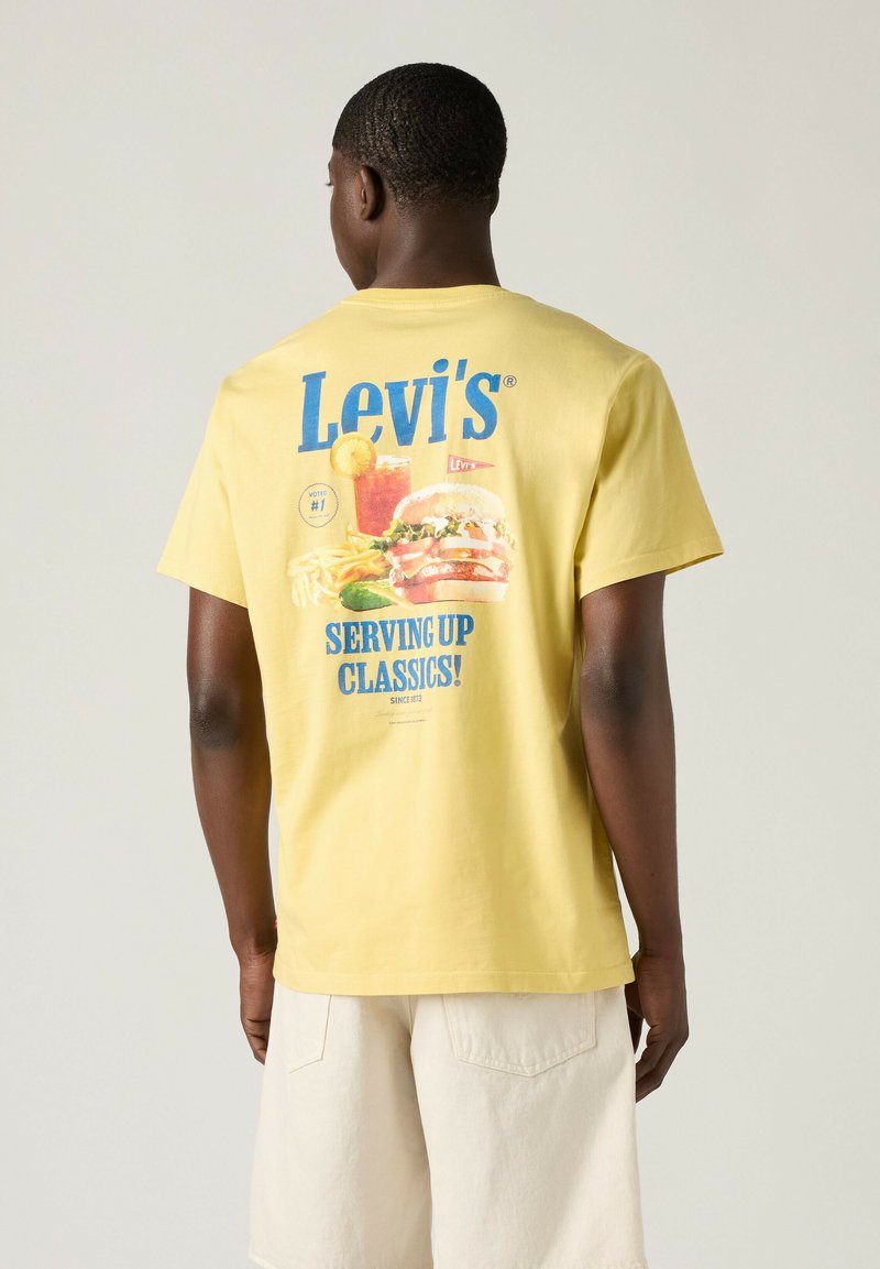 Ein Mann mit dem Rücken zur Kamera trägt ein gelbes Levi's T-Shirt mit Grafik von Burger, Pommes und Getränk sowie blauem Schriftzug „Levi's Serving Up Classics!“