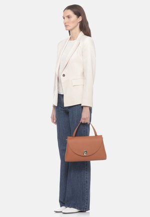 Donna con blazer color crema, top bianco, jeans blu a gamba larga, scarpe bianche, che tiene una borsa a mano in pelle marrone con chiusura dorata.