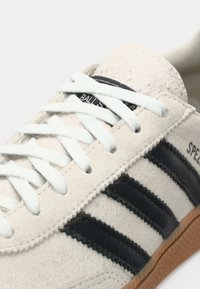 Beige mokkanahkainen tennari, jossa valkoiset nauhat. Mustat nahkasuikaleet ja kuvioitu purukumipohja. Brändäys näkyvissä sivussa.