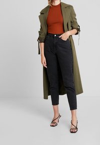 Kvinna som bär en olivgrön trenchcoat, en rostfärgad ribbad topp, högmidjade svarta jeans och svarta remsandaler, stående framför en enhetlig bakgrund.