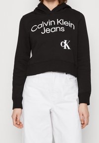 Sudadera corta negra con la inscripción "Calvin Klein Jeans" en blanco y un pequeño logo "ck" blanco en la parte inferior izquierda. Material de algodón, ajuste holgado.