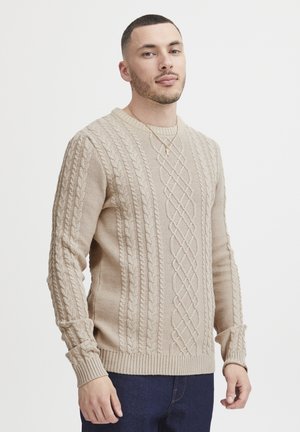 Beige cable-knit sweater featuring a textured pattern, round neckline, and ribbed cuffs and hem. Worn with blue denim jeans.