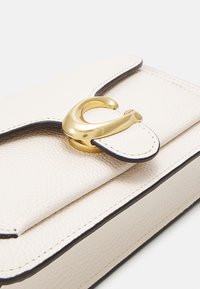 Bolso de cuero en color blanco roto con superficie texturizada, que presenta un broche de metal dorado y bordes negros. Solapa de cierre con un detalle de herrajes distintivo.