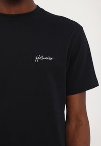 Fekete pamut póló rövid ujjakkal, fehér hímzett logóval a bal mellen. Klasszikus kerek nyakú és egyenes szabású.