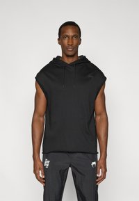 Μπλούζα χωρίς μανίκια με κουκούλα σε μαύρο χρώμα από μαλακό υλικό, με διακριτικό λογότυπο στο αριστερό στήθος. Συνδυασμένη με υφασμάτινο σκούρο παντελόνι με υφή.