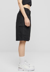 Sorte cargo shorts med midjebånd, som har to sidelommer og et kne-langt snitt, iført hvite joggesko og sokker.