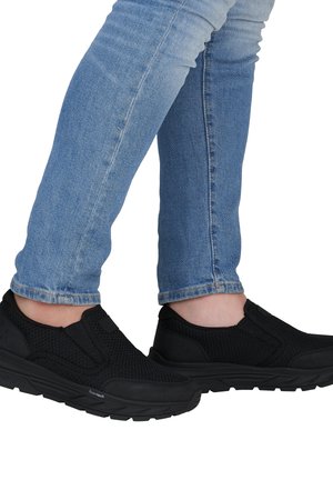 Person trägt hellblaue Jeans und schwarze Slip-On-Sportschuhe mit strukturierten Sohlen und Schaumpolsterung.
