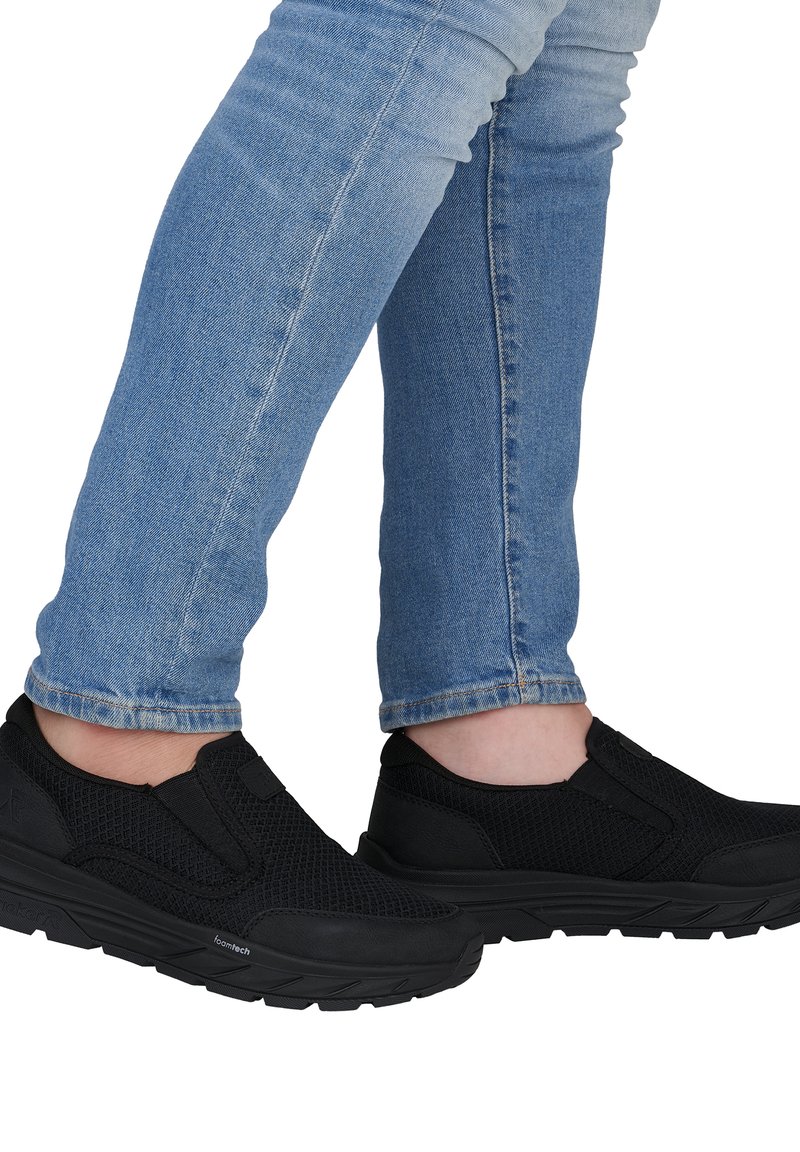 Person trägt hellblaue Jeans und schwarze Slip-On-Sportschuhe mit strukturierten Sohlen und Schaumpolsterung.