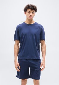 Νέος άνδρας με κοντά σγουρά μαλλιά, που φοράει μπλε κοντομάνικο πουκάμισο και αντίστοιχα σορτς, στέκεται μπροστά σε ένα απλό φωτεινό φόντο.