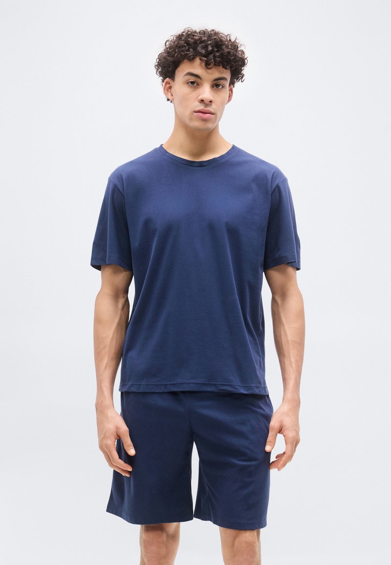 Νέος άνδρας με κοντά σγουρά μαλλιά, που φοράει μπλε κοντομάνικο πουκάμισο και αντίστοιχα σορτς, στέκεται μπροστά σε ένα απλό φωτεινό φόντο.