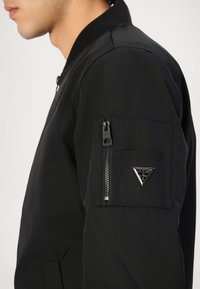 Black bomber jacket with a smooth texture, featuring a zippered pocket on the sleeve and a silver triangular logo accent.