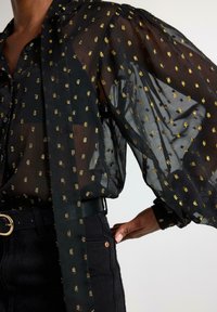 Sort transparent bluse med guldprikker, der har puffede ærmer og et bundet kravedesign, parret med højtaljede sorte jeans og et bælte.