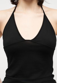 Žena oblečená v priliehavom čiernom halter top s hlbokým výstrihom do V, ktorý odhaľuje ramená a hornú časť hrudníka, na jednoduchom pozadí.