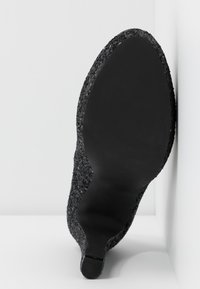 Semelle d'une chaussure noire à talon haut appuyée contre un mur blanc, montrant la semelle lisse et la tige en tissu texturé.