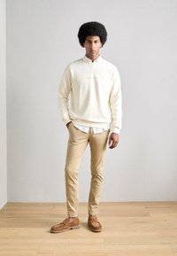 Licht crème sweatshirt met een verhoogde halslijn en klein logo, gecombineerd met beige slim-fit broek en bruine loafers, staand op een houten vloer.