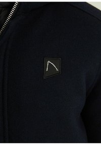 Jūras spēku zip-up jaka, izgatavota no teksturētas auduma, ar mazu melnu uzlīmi ar ģeometrisku baltu logotipu kreisajā krūtīs.