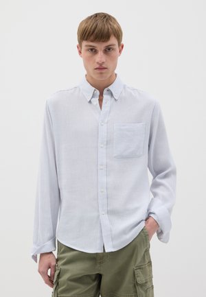 Camicia a maniche lunghe a righe azzurre e bianche con bottoni, tasca sul petto e orlo arrotondato, realizzata in tessuto morbido. Indossata con pantaloni cargo verde oliva.