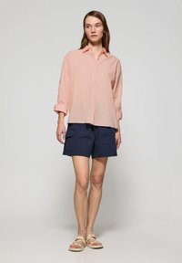 Camicia button-up rosa chiaro con maniche arrotolate, abbinata a pantaloni corti navy con tasche laterali e una vestibilità comoda. Sandali di colore chiaro.