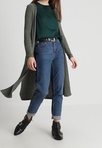 Cardigan verde a coste sopra una blusa teal, abbinato a jeans blu con risvolto e stivali neri alla caviglia con fibbie in metallo.