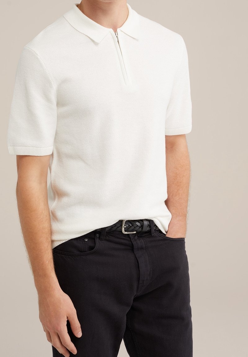 Polo blanc en tricot avec col zippé et manches courtes, associé à un jean noir en denim et une ceinture noire tressée.