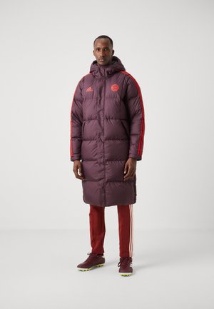 Abrigo puffer burdeos con capucha, que presenta franjas laterales rojas y un emblema. Combinado con pantalones deportivos rojos y zapatillas marrones con detalles amarillos.