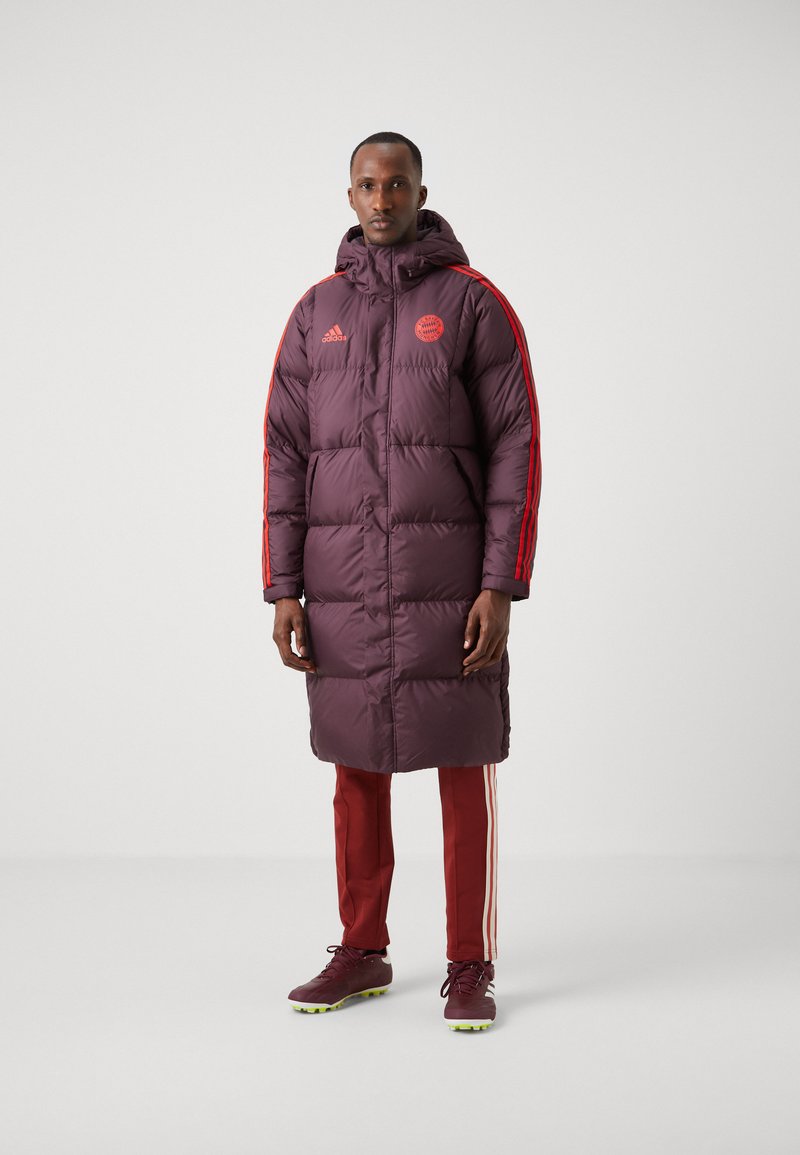 Abrigo puffer burdeos con capucha, que presenta franjas laterales rojas y un emblema. Combinado con pantalones deportivos rojos y zapatillas marrones con detalles amarillos.