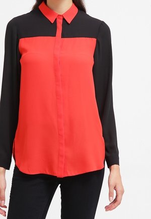 Nainen, jolla on päällään pitkähihainen pusero, jossa on musta yläosa ja hihat sekä kirkkaanpunainen etuosa ja kaulus, yhdistettynä mustiin housuihin.