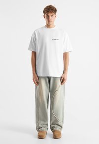 Vit bomull t-shirt med svart text "don't waste culture", kombinerad med ljusblå denimbyxor med en vågig söm och bruna slip-on skor.