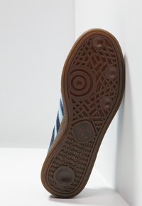 Suela de zapato atlético azul con goma marrón texturizada, diseñada con patrones para ofrecer agarre; presenta el logo de Adidas y elementos circulares integrados en el diseño.