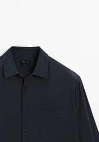 Camicia button-up blu scuro con colletto classico, maniche lunghe e taschino sul petto. Realizzata in tessuto testurizzato, presenta cinque bottoni.