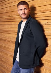 Dunkelblaues Blazer mit glatter Textur, Ein-Knopf-Verschluss und einer Fronttasche, getragen über einem weißen T-Shirt und kombiniert mit Denim.