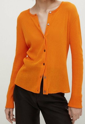 Femme portant un cardigan côtelé orange ajusté à boutons avec manches longues et un pantalon noir, debout devant un fond uni.