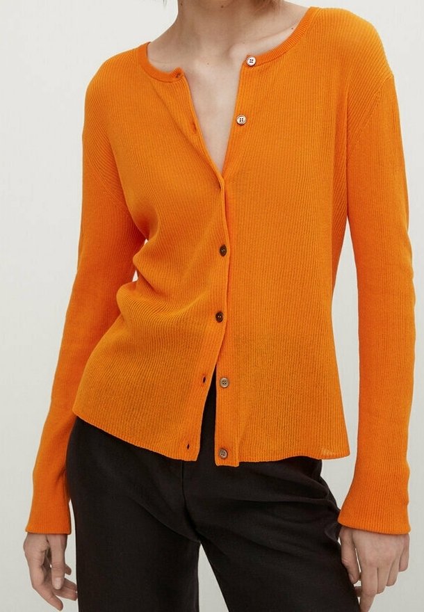 Femme portant un cardigan côtelé orange ajusté à boutons avec manches longues et un pantalon noir, debout devant un fond uni.